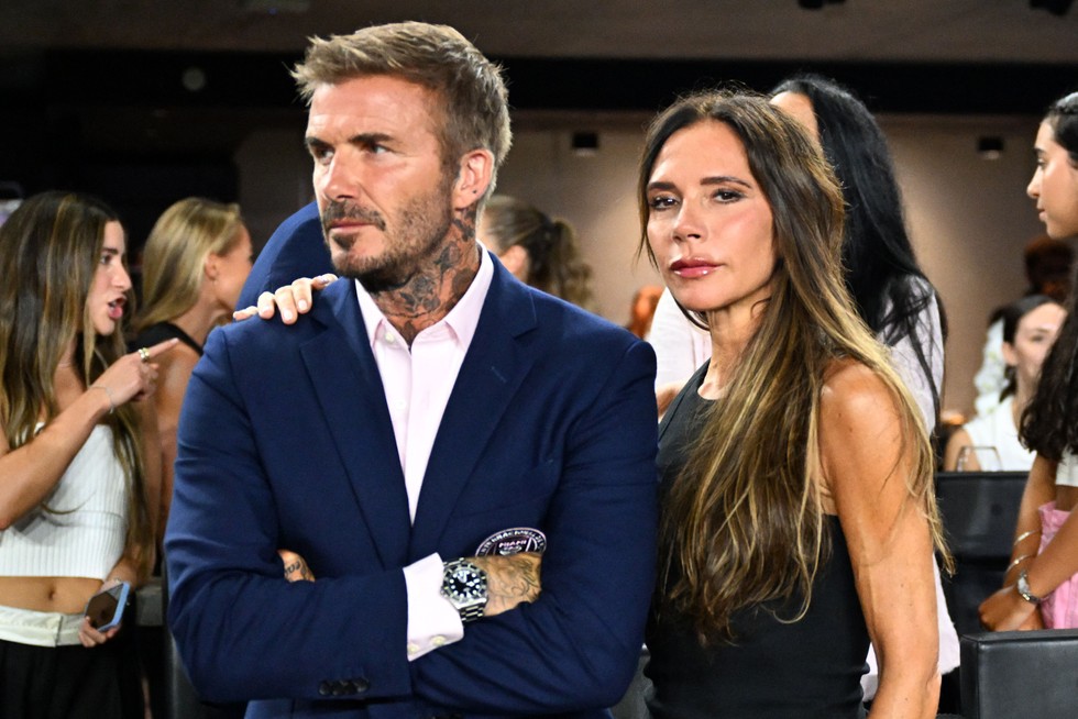 David Beckham and Victoria Beckham. David has his arms folded looking off to the side and Victoria has her arm around his shoulder looking to camera.