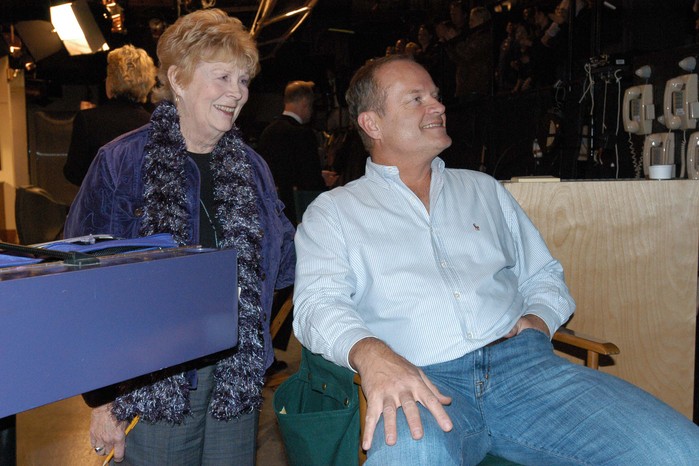 Kelsey Grammer is seated in a director's chair, with Gabrielle James standing close behind him, both are smiling and looking to their left-hand side