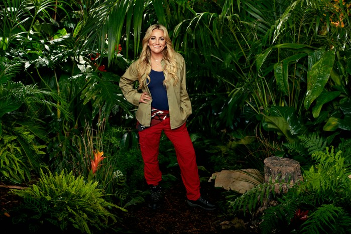 Jamie Lynn Spears wearing her I'm a Celebrity camp clothes (khaki shirt, blue vest and red trousers) while posing in front of some bushes ahead of I'm a Celebrity 2023