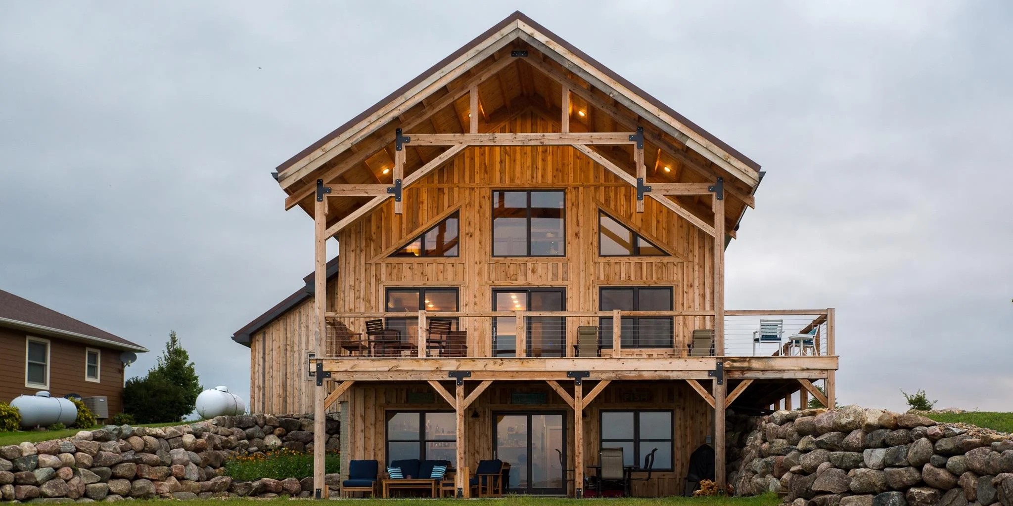 exterior view of lakeside SIP home; deck, balcony, and roof overhang have geometric post-and-beam constructions; knotty wood siding; low stone wall surrounds - photo