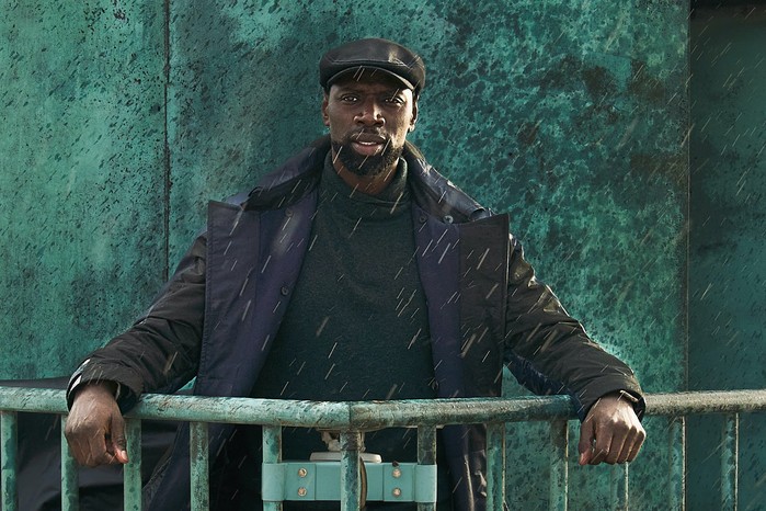 Assane Diop in Lupin wearing a dark jacket and cap, leaning on a rail