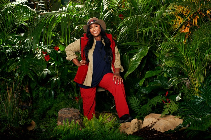 I'm a Celebrity star Nella Rose dressed in her camp gear (red trousers, khaki shirt, red gilet and navy top) while posing in front of some bushes ahead of the 2023 show.