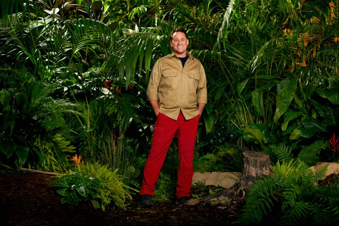 Nick Pickard dressed in a khaki shirt and red trousers while posing in front of some trees for I'm a Celebrity 2023.