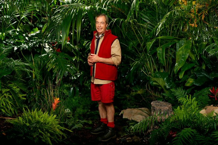 Nigel Farage wearing his I'm a Celebrity camp clothing (khaki shirt, red gilet, red trousers and black boots) as he poses in front of some bushes ahead of I'm a Celebrity 2023.