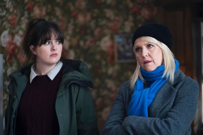 Alison O'Donnell as Tosh in Shetland, standing next to Ashley Jensen as Ruth Calder. They're stood inside a house, with Ruth looking straight ahead with her arms crossed, a stern expression on her face, and Tosh looking at her with a concerned expression on her face