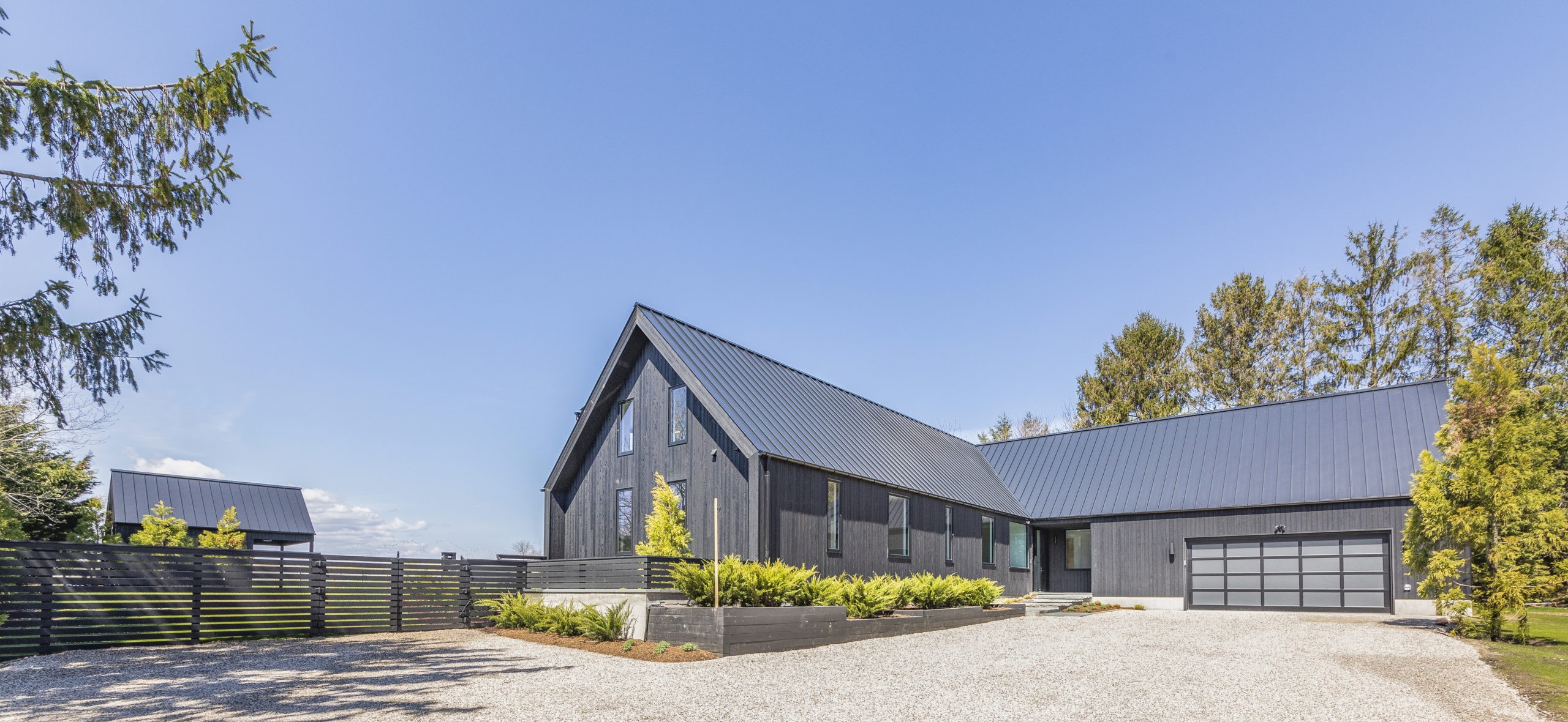 exterior view home in rural setting; broad gravel drive in foreground; home has gray siding and metal roof with arrached garage; landscaping, outbuilding and expansive blue sky in background - photo